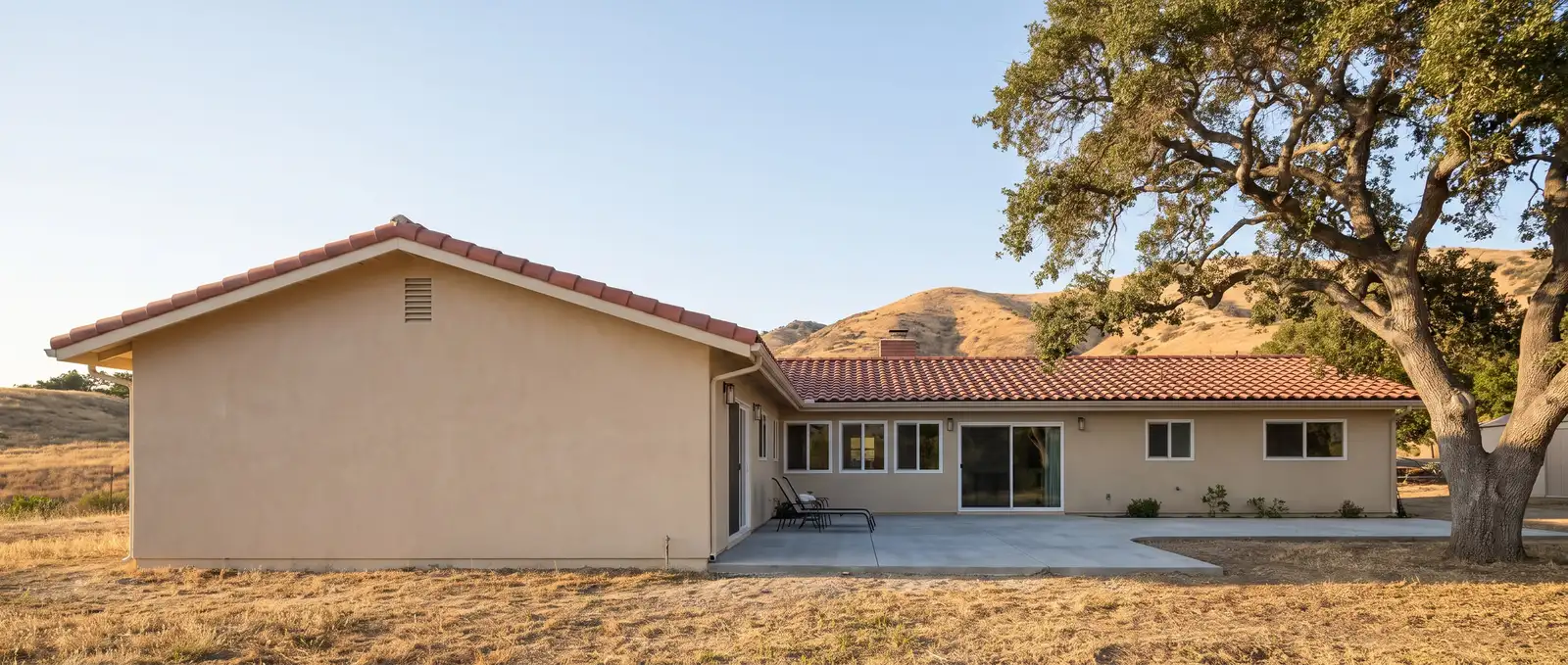 Casa estilo rancho de un piso con una ampliación trasera nueva en estuco y teja roja iguales a los de la casa, con cerros dorados y un encino detrás, ilustración generada con IA